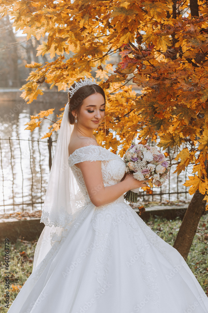 A beautiful bride stands in nature near the autumn leaves of trees with a bouquet of flowers. Close-up wedding portrait of a young bride. Wedding photography.