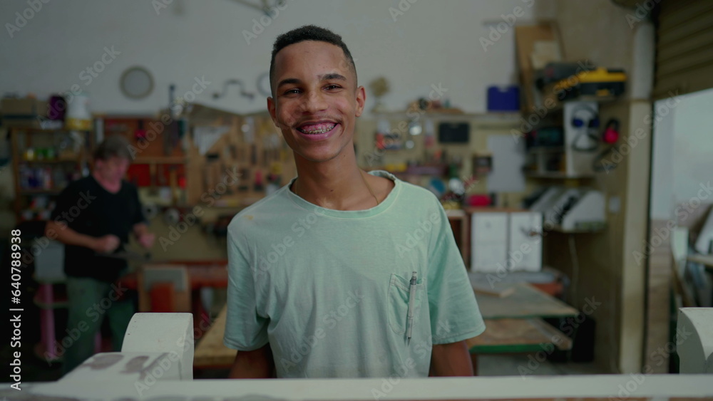 Fototapeta premium Portrait of a happy black Brazilian employee of carpentry workshop, local small business occupation. Young apprentice looking at camera smiling