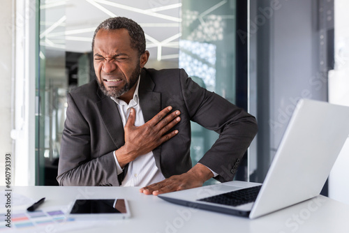 Heart attack in mature experienced man inside office at workplace, senior african american holding hands on chest, pain is severe in boss in business suit. © Liubomir