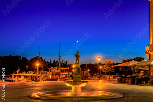 The fountain boy with the fish on main square in Rovinj, Croatia at the harbor at night with waning moon