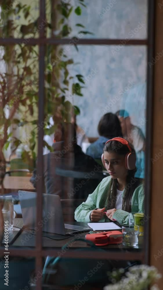 With headphones on, a pretty young lady is working on her laptop in a café. With headphones on, a pretty young lady is working on her laptop in a café.