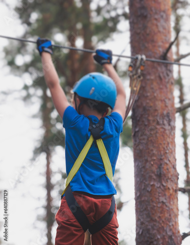 View of high ropes course, kids of climbing in amusement acitivity rope park, passing obstacles and zip line on heights, children teenagers in equipment gear between the trees on heights, summer day