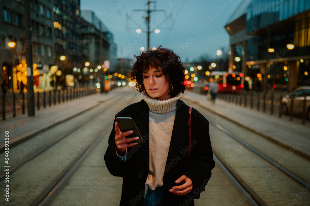 Fototapeta premium Dark haired woman standing at the train station and looking down to her phone.