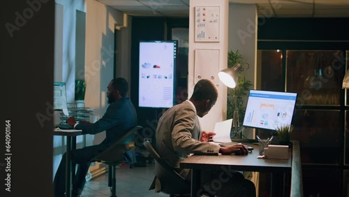 Revealing shot of employees checking company figures, postings and documents for accuracy during nighshift. Staff member working overnight, analyzing stored computerized financial information