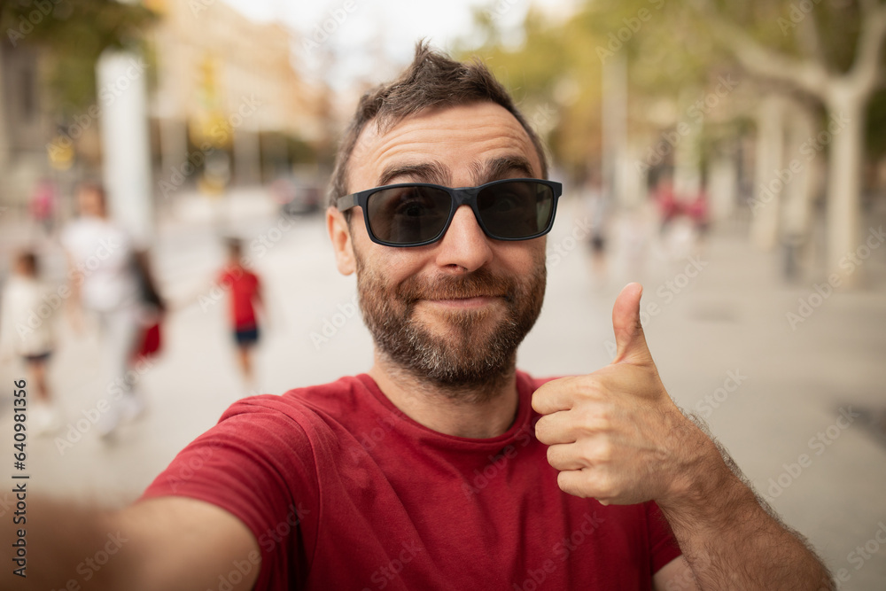 A young, bearded man, wearing sunglasses, smiles at the camera, while taking a selfie with his cell phone during a walk in the city. A young, bearded man, wearing sunglasses, smiles at the camera, while taking a selfie with his cell phone during a walk in the city.
