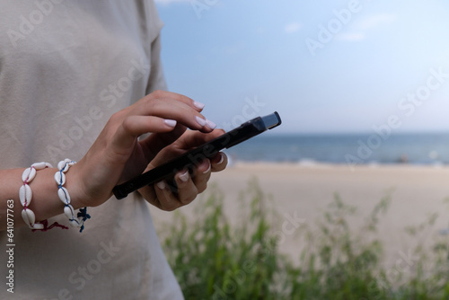 Female hand with shell bracelets holding smartphone. Unrecognizable woman using mobile phone on the beach seacoast. Holiday vacation advertisement concept. Social media 