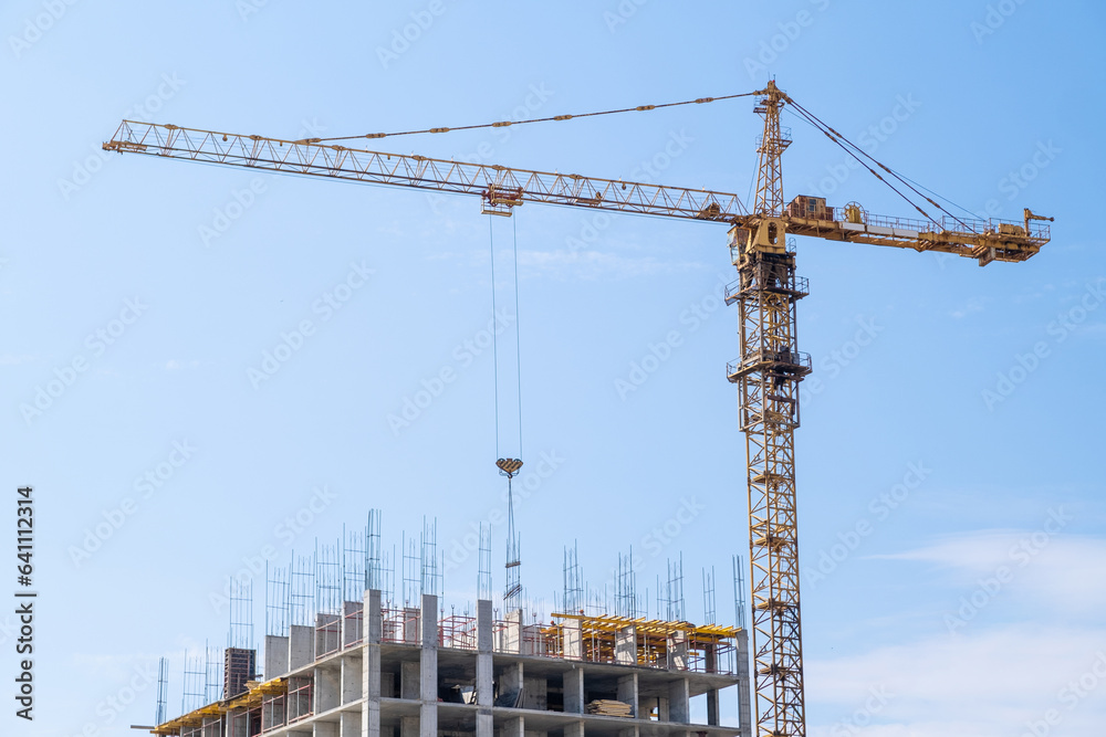 A crane and a building under construction against a blue sky background. Builders work on large construction sites, and there are many cranes working in the field of new construction. A crane and a building under construction against a blue sky background. Builders work on large construction sites, and there are many cranes working in the field of new construction.