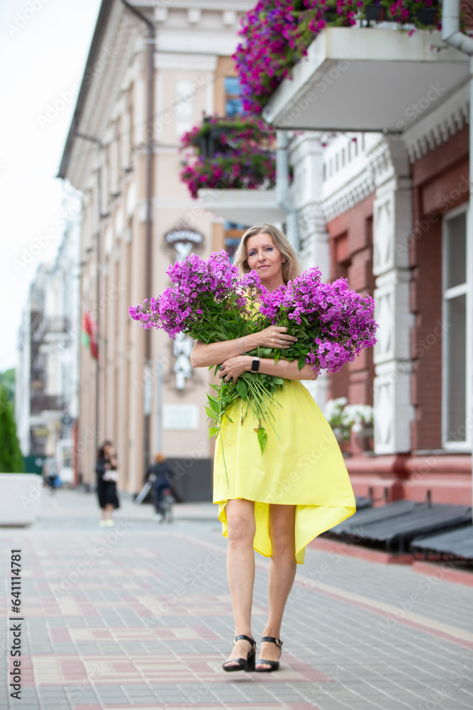 A beautiful elderly woman with a bouquet of flowers walks through the streets of the city. Age model blonde in full growth. A woman in her fifties in a beautiful dress.