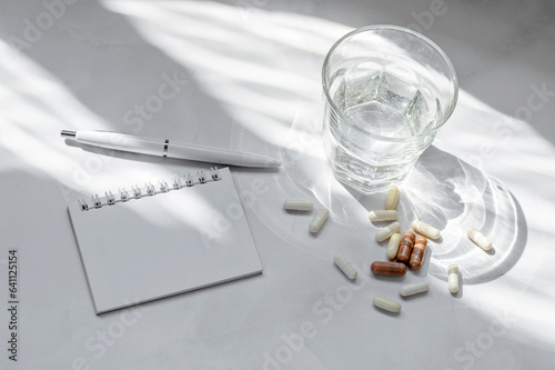 Pills and capsules, glass with pure water, blank notebook mockup, on white table background with aesthetic sunlight shadow. Healthy morning routine planning, beauty, nutrition supplement and vitamins