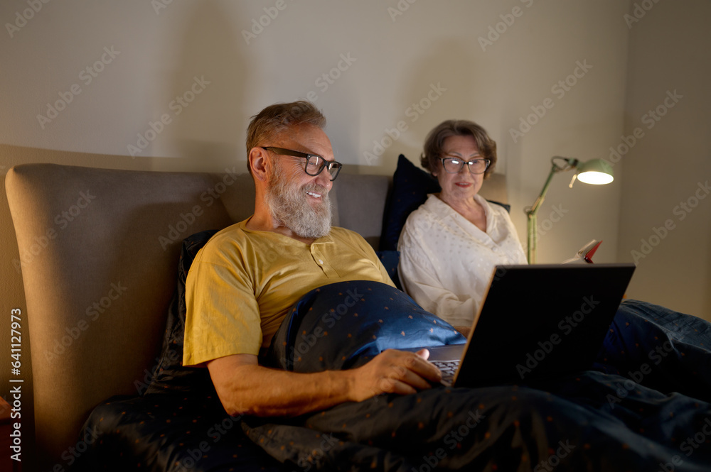 Senior couple using laptop lying on bed in bedroom at comfortable home Senior couple using laptop lying on bed in bedroom at comfortable home