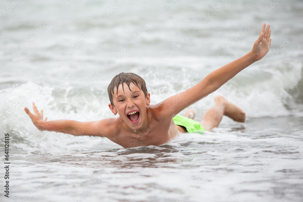 A cheerful happy boy plays with sea waves, spreads his arms and screams with delight.