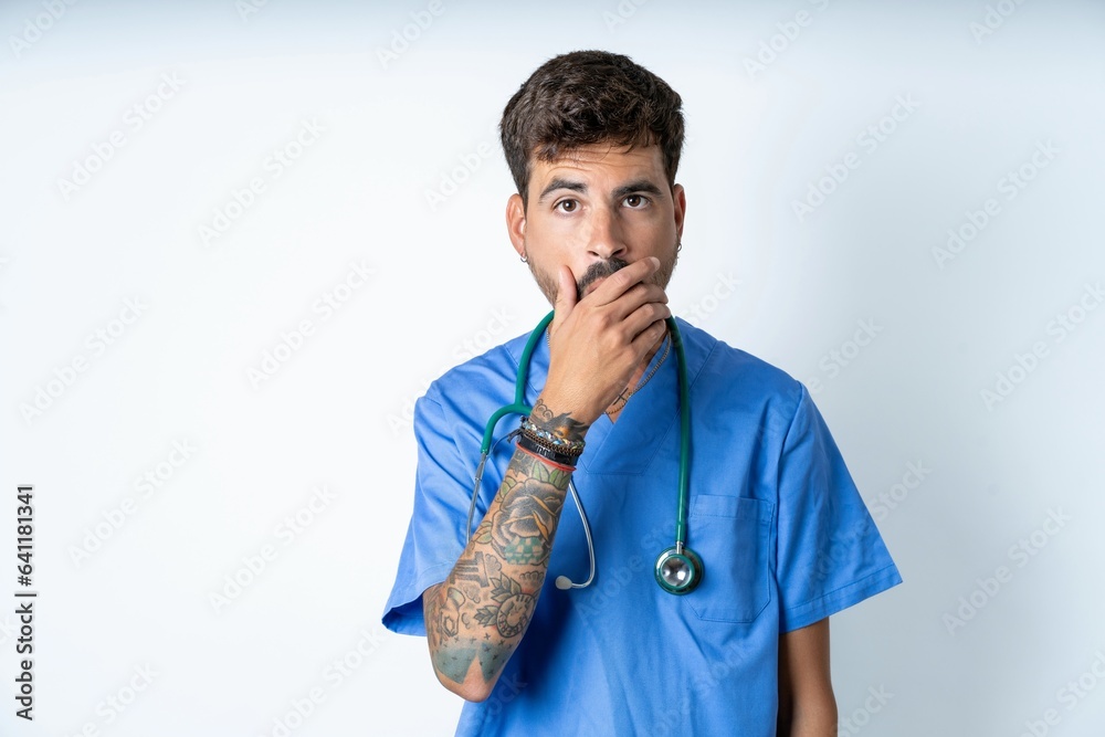 Emotional young caucasian doctor man wearing blue medical uniform gasps from astonishment, covers opened mouth with palm, looks shocked at camera. Emotional young caucasian doctor man wearing blue medical uniform gasps from astonishment, covers opened mouth with palm, looks shocked at camera.