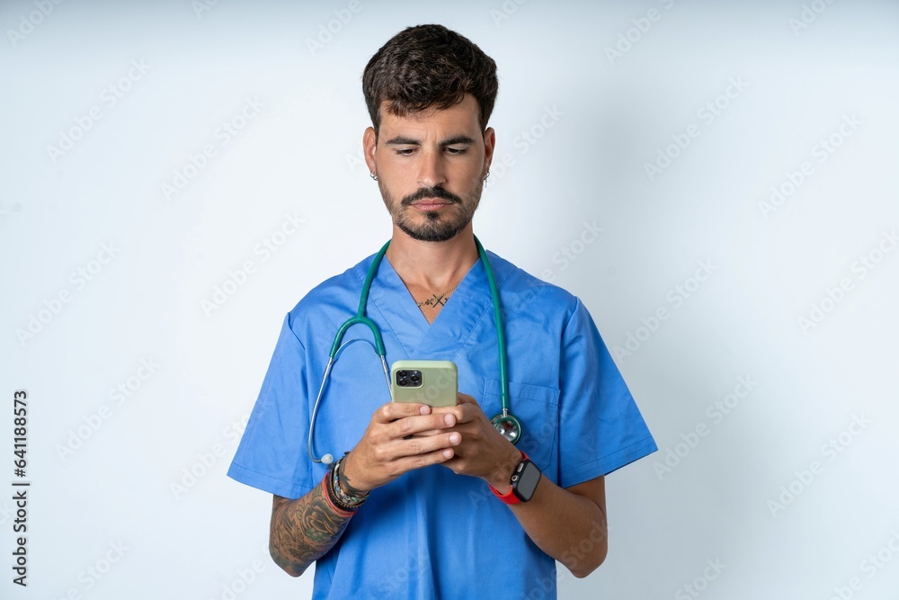 Excited young caucasian doctor man wearing blue medical uniform winking and eye hold smart phone use read social network news Excited young caucasian doctor man wearing blue medical uniform winking and eye hold smart phone use read social network news