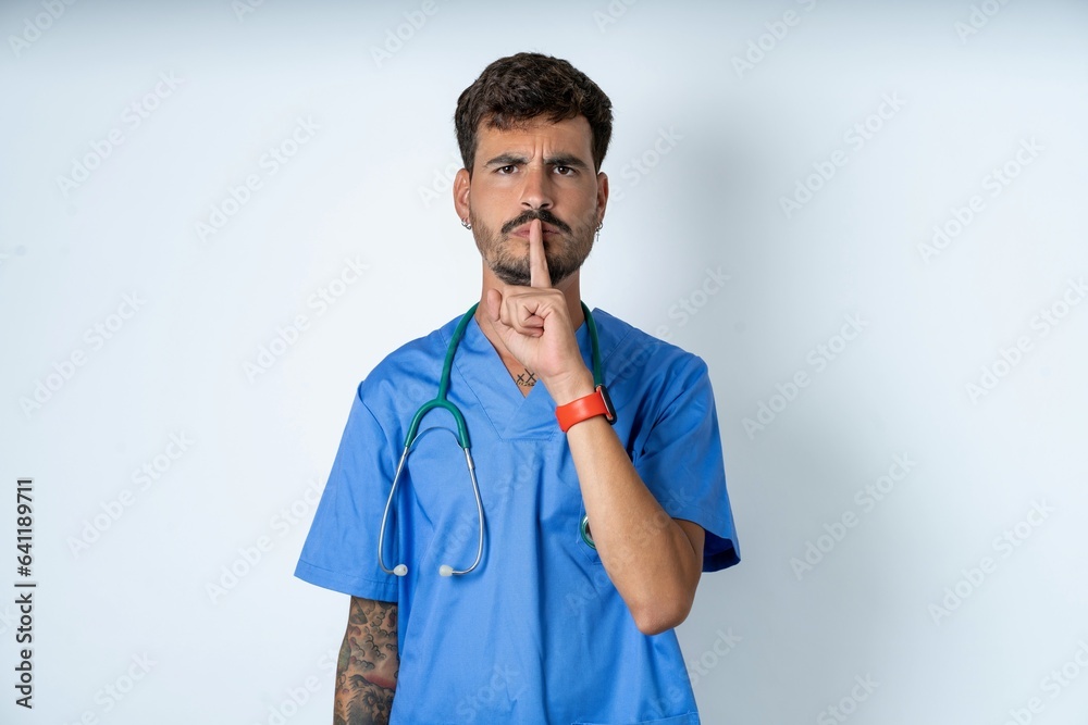 young caucasian doctor man wearing blue medical uniform silence gesture keeps index finger to lips makes hush sign. Asks not to share secret young caucasian doctor man wearing blue medical uniform silence gesture keeps index finger to lips makes hush sign. Asks not to share secret