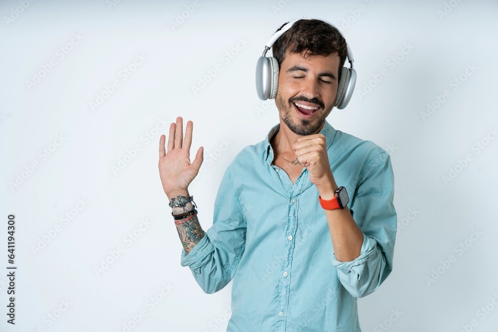 Happy young caucasian man wearing green shirt sings favourite song keeps hand near mouth as if microphone wears wireless headphones, listens music Happy young caucasian man wearing green shirt sings favourite song keeps hand near mouth as if microphone wears wireless headphones, listens music
