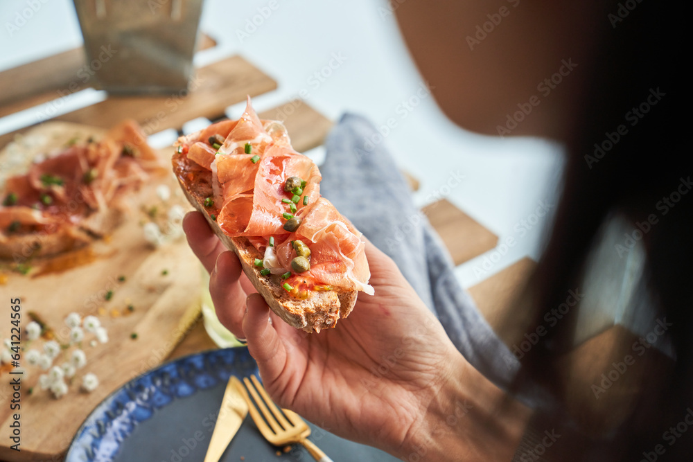 Crop woman enjoying delicious toast with prosciutto on terrace