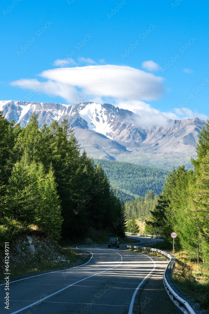 Scenic view from sunlight grassy hill with road to high mountain range. Beautiful sunny mountain landscape with large snow peak mountains. Road trip to Altai. Scenic view from sunlight grassy hill with road to high mountain range. Beautiful sunny mountain landscape with large snow peak mountains. Road trip to Altai.