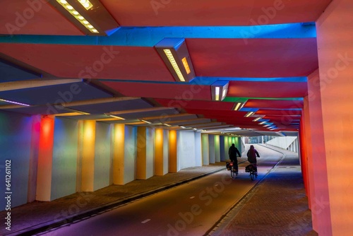 Two cyclists ride side by side through a colorfully lit bicycle tunnel