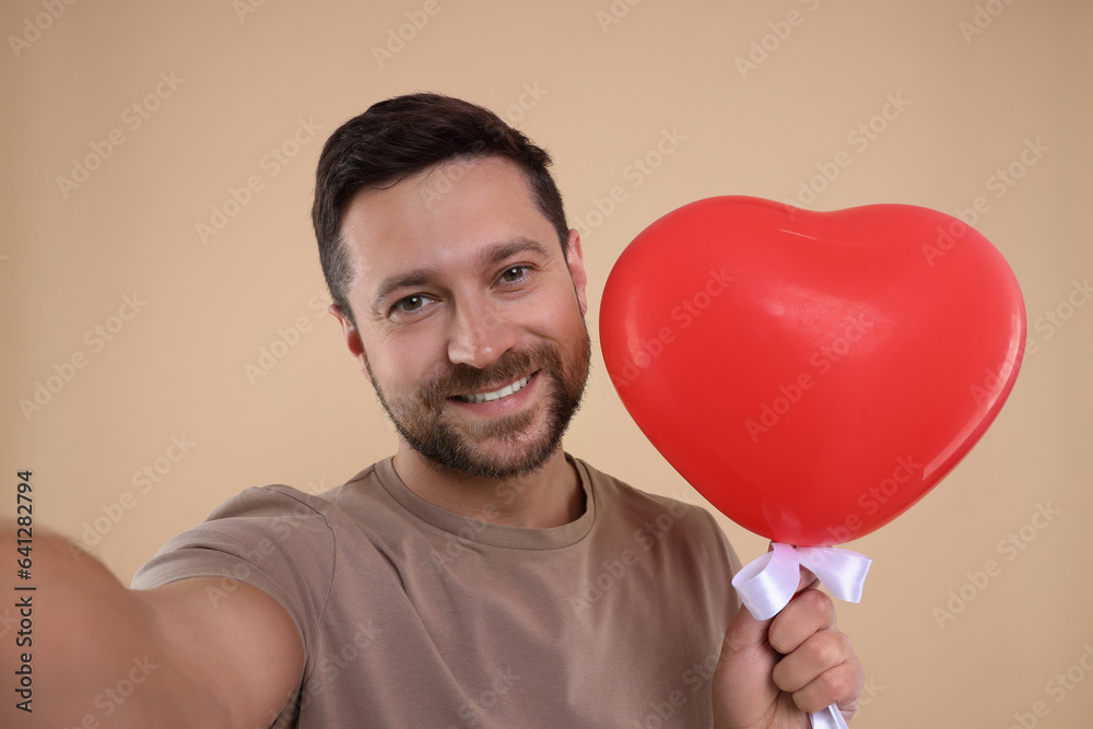 Man holding red heart shaped balloon and taking selfie on beige background Man holding red heart shaped balloon and taking selfie on beige background