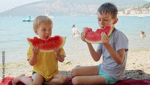 Two cute boys sitting on the sea beach towel and eting sweet watermelon. Summertime, holiday, vacation, happiness and relaxation photo