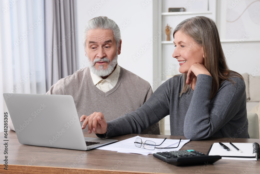 Elderly couple with papers and laptop discussing pension plan at wooden table in room