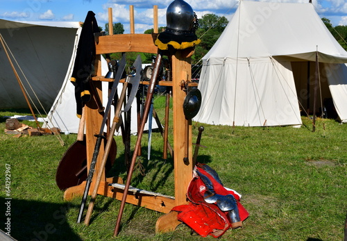A close up on a wooden stand with various medieval weapons displayed on it, including some wooden swords, polearms, shields, axes, scimitars, and other ones seen on a sunny summer day in Poland