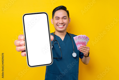 Cheerful young Asian male doctor or nurse wearing blue uniform holding cash money and showing a mobile phone blank screen isolated on yellow background. Healthcare medicine concept