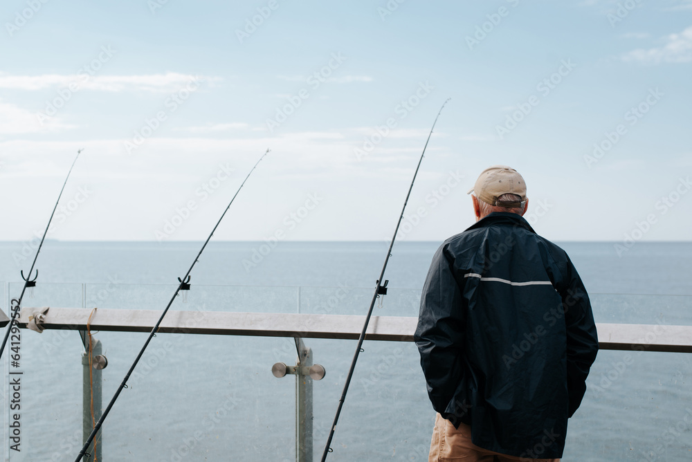 Old fisherman with fishing rods fishing on bridge looking at horizon of sea, back view