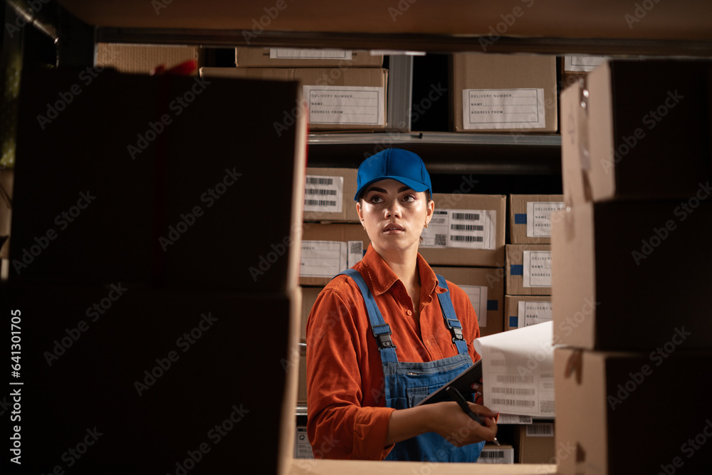 Delivery woman in blue overalls with clipboard and pen over warehouse background. Profession, job and people concept Delivery woman in blue overalls with clipboard and pen over warehouse background. Profession, job and people concept