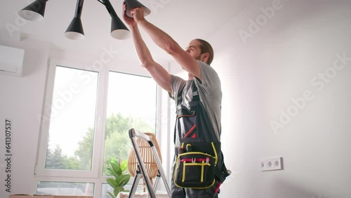 Electrician man screwing out light bulb from chandelier. Craftsman standing at ladder and changing light bulb. Electric specialist holding tools at lower back. Repairman checking light bulb.