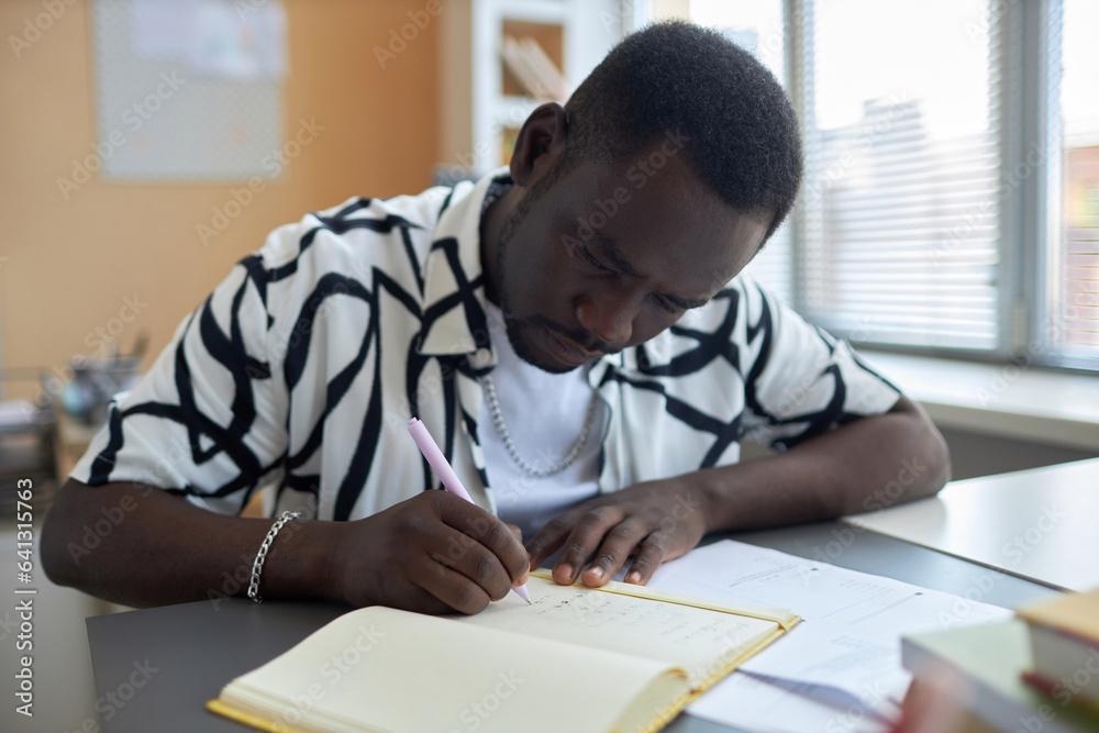 Serious African American male student bending over open copybook while sitting by desk at lesson and writing down answers to test questions Serious African American male student bending over open copybook while sitting by desk at lesson and writing down answers to test questions