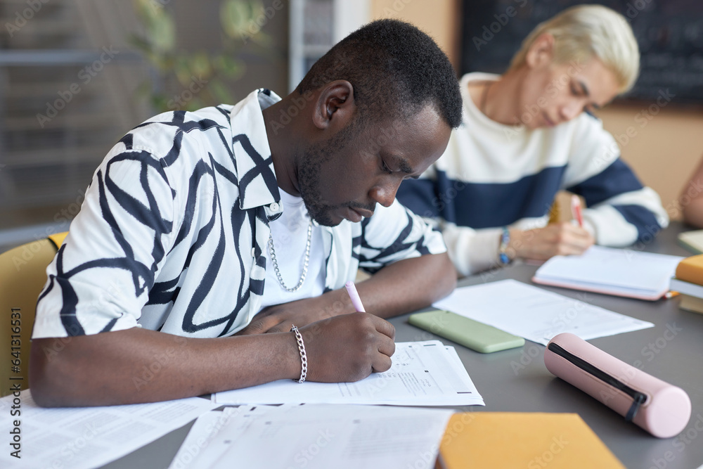 Serious African American guy making notes or putting ticks on paper while answering questions of examination test against Asian classmate Serious African American guy making notes or putting ticks on paper while answering questions of examination test against Asian classmate
