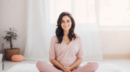 Portrait of a happy pregnant woman sitting cross legged at home. Expecting mother with folded legs stroking her belly smiling and looking at the camera on a sunny day. Young pregnant woman with smile. © Valua Vitaly