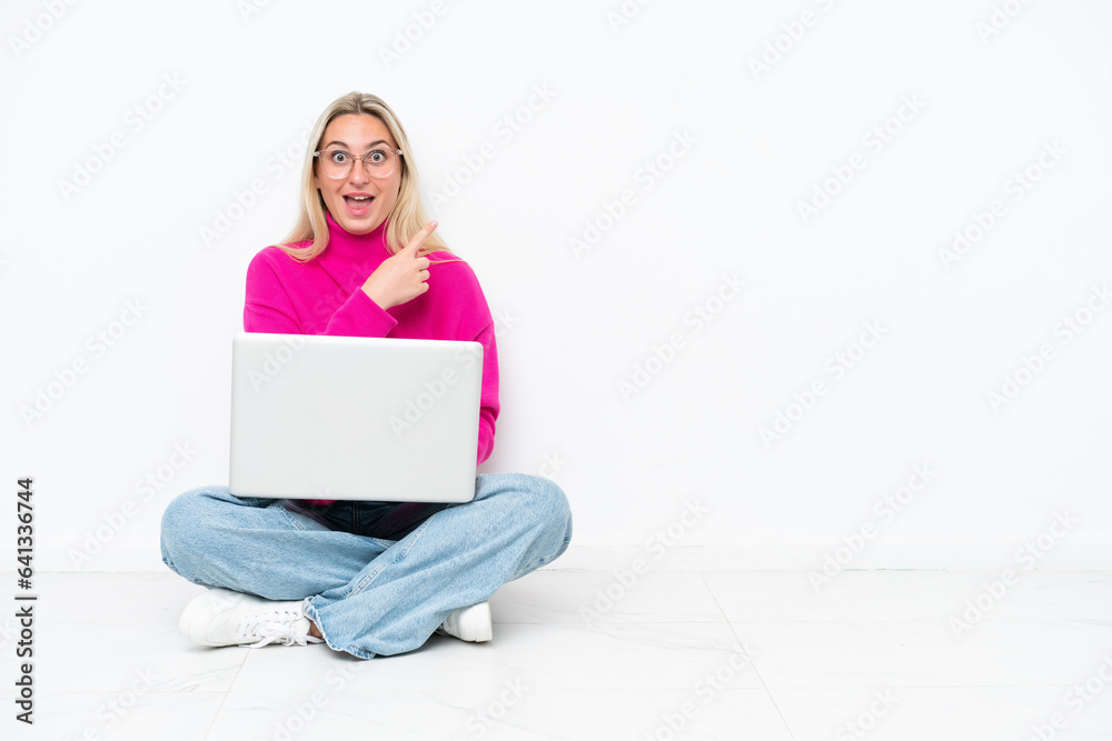 Fototapeta premium Young caucasian woman with laptop sitting on the floor surprised and pointing side