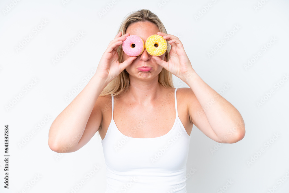 Young caucasian woman isolated on white background holding donuts in eyes with sad expression