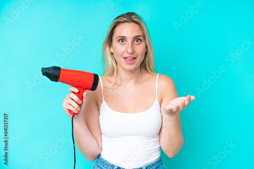 Young caucasian woman holding a hairdryer isolated on blue background with shocked facial expression
