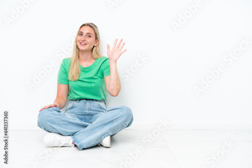 Young caucasian woman sitting on the floor isolated on white background saluting with hand with happy expression