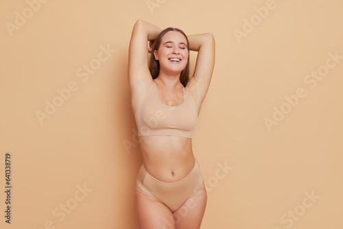 Slim female model in body-colour sports underclothing poses with hands over her head inside studio, gentle smile and closed eyes, best underwear concept, copy space photo