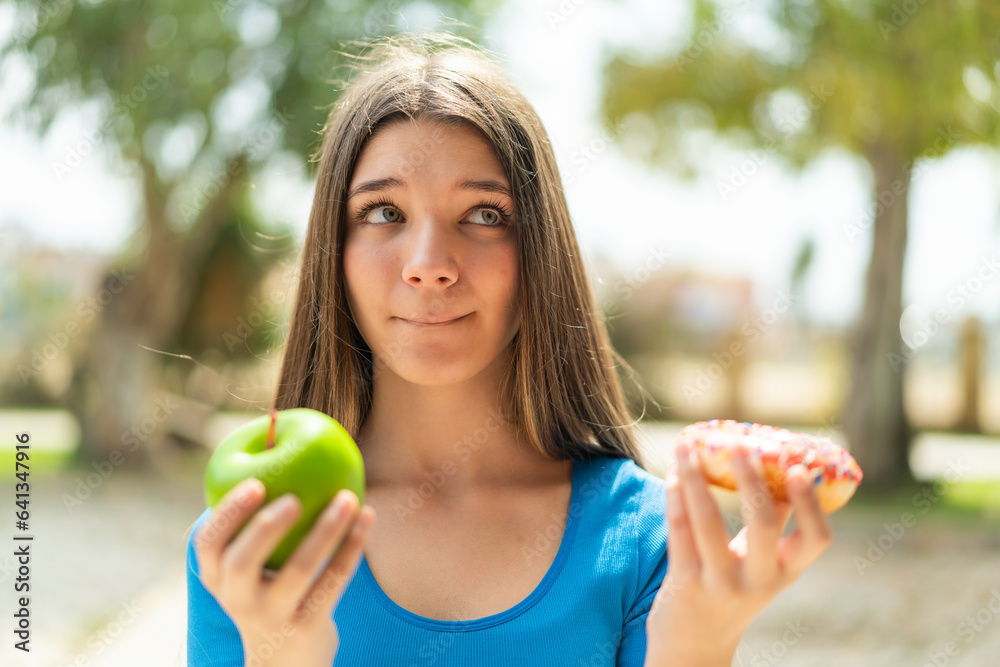 Teenager girl at outdoors holding apple and donut and having doubts Teenager girl at outdoors holding apple and donut and having doubts