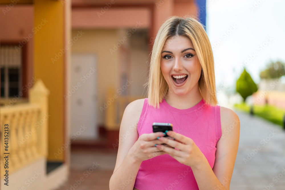 Young pretty blonde woman at outdoors surprised and sending a message Young pretty blonde woman at outdoors surprised and sending a message
