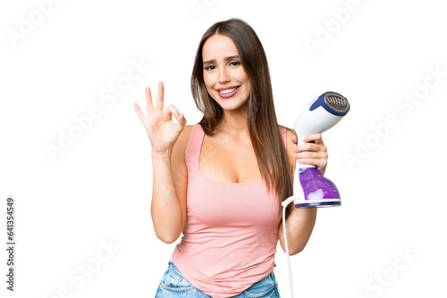 Young beautiful woman holding a vertical steam iron over isolated chroma key background showing ok sign with fingers
