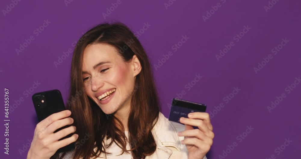 Young woman wear white jeans jacket using mobile cell phone hold in hand credit bank card do online shopping book tour isolated on purple background. Wow surprised girl look at phone, show credit card Young woman wear white jeans jacket using mobile cell phone hold in hand credit bank card do online shopping book tour isolated on purple background. Wow surprised girl look at phone, show credit card