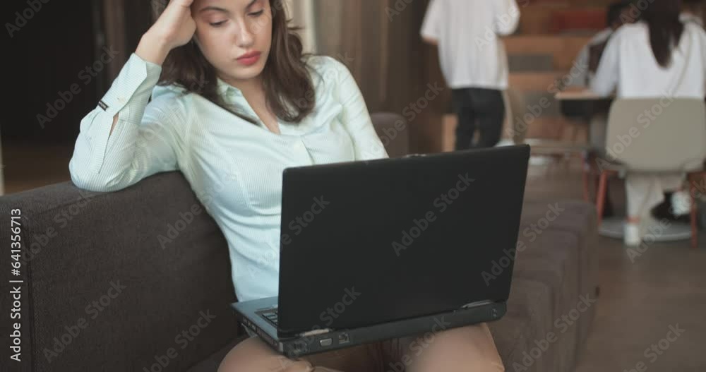Young female freelancer thinking how to solve a problem while sitting in the office sofa, coworkers blurred in the back, camera tilt Young female freelancer thinking how to solve a problem while sitting in the office sofa, coworkers blurred in the back, camera tilt