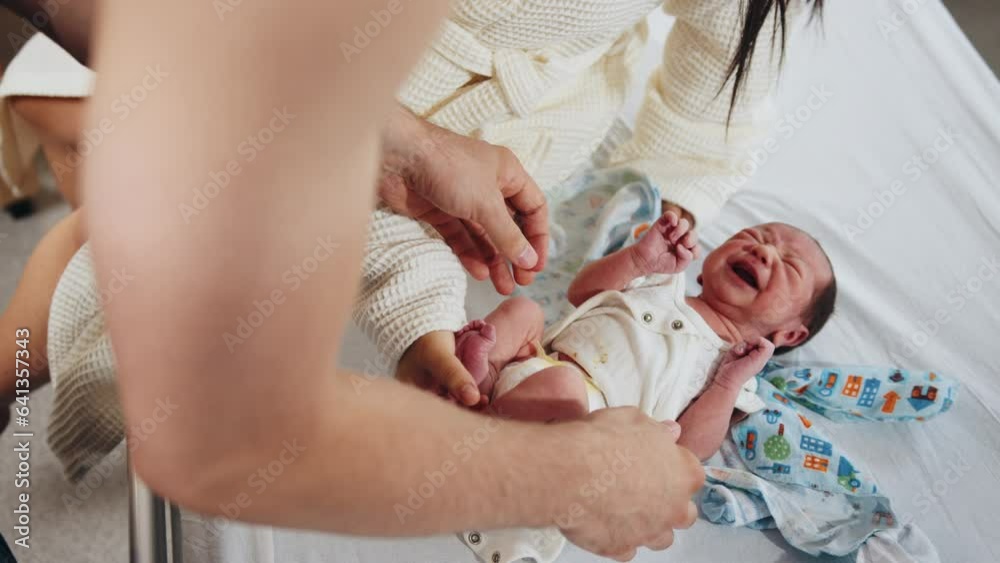 Multiethnic couple holding and admiring their newborn baby in hospital in post natal department. Unknown mother taking care of her baby boy or girl putting on clothes - newborn infant lay down on bed
