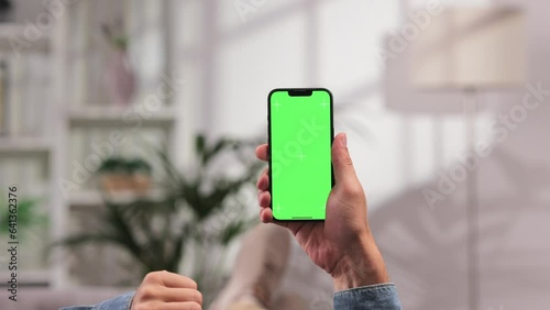 Caucasian man scrolls sideways and taps the green screen of a smartphone with his thumb, lying on the sofa in the afternoon in a bright living room. Close up of a male hand holding a mobile phone.