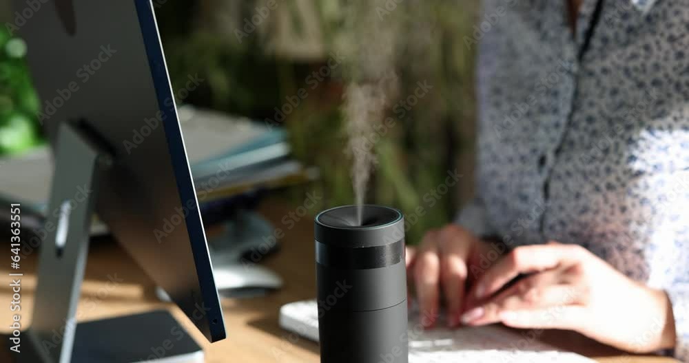 Woman is typing on computer keyboard and diffuser diffuses fresh scent. Female worker enjoying aroma of essential oils in office