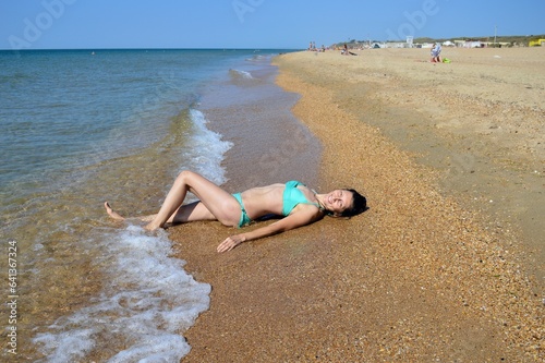 A girl in a turquoise swimsuit lies on the sand by the seashore.