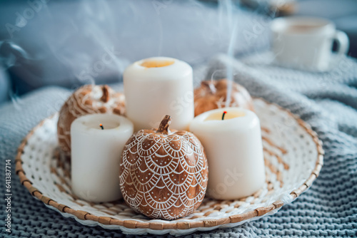 Autumn fall cozy mood composition for hygge home decor. Orange pumpkins decorated with mandalas, white blown candles with smoke on wicker plate on the knitted gray plaid. Selective focus. Copy space