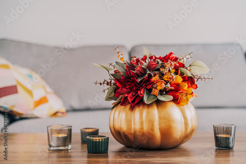 Fototapeta Naklejka Na Ścianę i Meble -  Autumn bouquet of bright artificial flowers in a golden pumpkin vase, burning candles on the wooden coffee table in living room interior with sofa. Hygge home fall decor for a cozy home atmosphere.