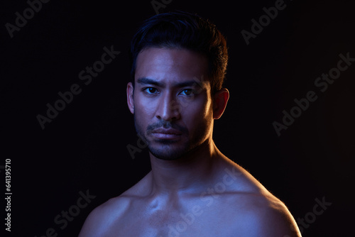 Blue light, man and portrait with creative, art and dark background in studio. Sexy, serious face and male model from Indonesia with skin glow, grooming care and wellness for skincare treatment © Joanrae P/peopleimages.com